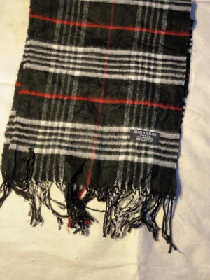 Rutig halsduk i kashmir från Burberry - Snygg halsduk från Burberry i 100% kashmir. Klassisk svart, vit och röd rutig design med fransar i ändarna. Tillverkad i England och supermjuk, perfekt för att lyfta din outfit med en tidlös accessoar.