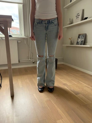 Ljusblå slitna bootcut jeans - Säljer ett par ljusblå bootcut jeans low waist med slitningar vid knäna och broderade detaljer på bakfickorna. Jeansen är avklippt ner till men passar mig som är 163 cm. 