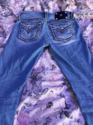 Ljusblå Miss Me jeans med broderi - Säljer ett par ljusblå jeans från Miss Me med snygga kontrastsömmar och detaljerade bakfickor med broderi och knappar. Jeansen har en klassisk femficksmodell och en metallplatta med M-logga bak i midjan. Perfekta för dig som gillar statement-jeans!