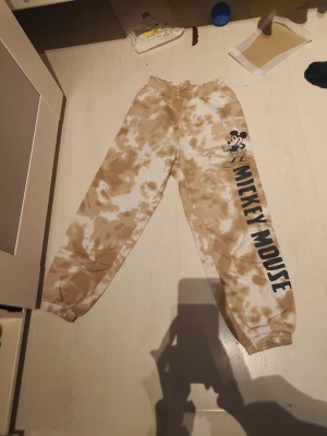 Beige tie-dye mjukisbyxor Mickey Mouse - Säljer ett par beige och vita tie-dye mjukisbyxor från Disney med Mickey Mouse-tryck och text längs ena benet. Byxorna har resår i midjan och vid bensluten för en skön och avslappnad passform. Perfekta för chill dagar hemma eller när du vill ha en lekfull stil.