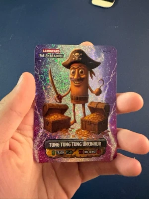 Sinnesjuk pull: Tung tung tung uncinhur - Säljer ett glittrigt samlarkort från Lamincard som jag själv skulle säga att är riktigt stilrent. Med motiv av en tecknad pirat med svärd och krokhand, samt två skattkistor fyllda med guld. Kortet har lila och silver bakgrund och texten 'TUNG TUNG TUNG UNCINIHUR' längst ner.