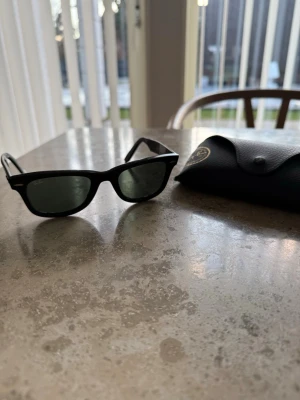 Svarta Ray-Ban Wayfarer solglasögon - Klassiska svarta Wayfarer solglasögon från Ray-Ban med gröntonade glas och robust plastbåge. Ikonisk design som passar till alla stilar. Kommer med originalfodral i svart läderlook.