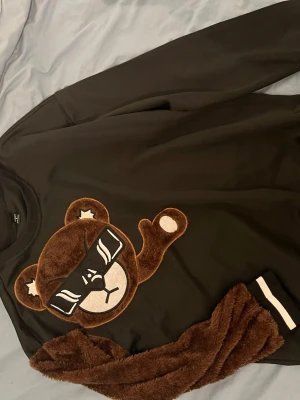 Svart sweatshirt med nalleapplikation - Cool svart sweatshirt med stor brun nalleapplikation framtill. Nallen har solglasögon och är gjord i mjuk fuskpäls. Tröjan har ribbade muddar med vita ränder vid ärmslut och nederkant. Perfekt för dig som gillar streetwear och unika detaljer.