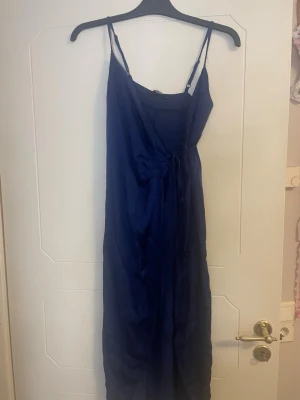 Marinblå långklänning Bik Bok XS - Snygg marinblå långklänning från Bik Bok i slätt, glansigt material. Klänningen har smala axelband, omlott-detalj framtill och knyt i midjan för en feminin siluett. Perfekt för sommarkvällar eller när du vill känna dig extra fin.