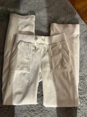 Vita mjukisbyxor från Juicy Couture - Säljer ett par vita mjukisbyxor från Juicy Couture med raka ben, bred resår i midjan och dragsko. Byxorna har två stora fickor fram och en liten broderad logga på ena fickan. Perfekta för chill dagar och riktigt sköna att ha på sig.