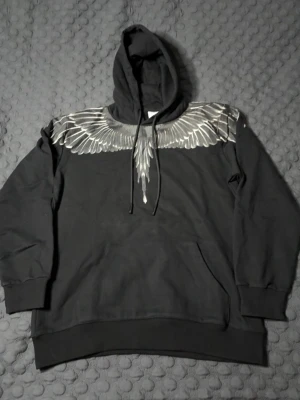 Svart hoodie från Marcelo Burlon  - Säljer en svart hoodie från Marcelo Burlon med ikoniskt vingeprint i vitt och grått över axlar och bröst, samt på ryggen. Hoodien har huva med dragsko och en stor magficka. Tillverkad i mjuk bomull för maximal komfort och streetwear-vibe. STORLEK M, PASSAR M. HELT NY ALDRIG ANVÄND.