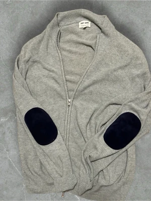 Soft goat 100% Kashmir - Snygg grå ziptröja från Soft Goat med svarta mockaliknande armbågslappar. Tröjan har ribbade muddar och hög krage. Perfekt för dig som gillar en stilren look med en twist. Materialet känns mjukt och skönt mot huden.