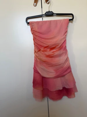  kortklänning med volang - Supersöt axelbandslös kortklänning i olika rosa nyanser med omlott-detalj och volang nedtill. Klänningen har en lätt rynkad effekt på sidan och är gjord i ett tunt, flowy material som känns perfekt för sommaren. Använd 1 gång! Passar mos som är S!