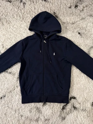 Polo Ralph Lauren Zip Hoodie - Snygg mörkblå hoodie från Polo Ralph Lauren med dragkedja framtill och klassisk logga broderad på bröstet. Hoodien har två fickor fram, justerbar huva med snören och är tillverkad i mjukt bomullsmaterial. Perfekt för en avslappnad och stilren look.
