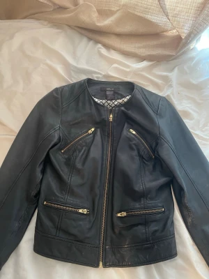 Svart skinnjacka från Ellos - Snygg svart skinnjacka från Ellos med fyra dragkedjefickor framtill och rund halsringning. Jackan har en stilren design med guldfärgade dragkedjor och figurnära passform. Perfekt för att lyfta vilken outfit som helst.