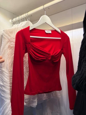 Röd långärmad topp från Oh Polly - Trendig röd topp med långa ärmar och markerad byst med draperade detaljer. Toppen har en tight passform och fyrkantig ringning som ger en modern look. Perfekt för dig som vill sticka ut med en färgstark och unik topp.