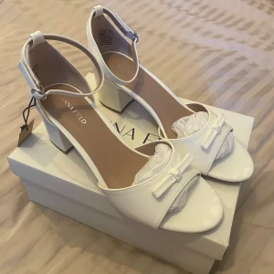 Vita sandaletter med blockklack Anna Field - Snygga vita sandaletter från Anna Field med öppen tå, bred rem framtill dekorerad med en liten rosett och pärla. Justerbart ankelband med spänne. Skorna har en stabil blockklack och är tillverkade i ett glansigt syntetmaterial som ger en fräsch look. Nypris 339kr (endast testade)