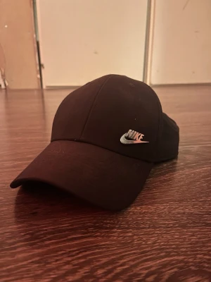 Svart Nike keps Dri-FIT M/L - Snygg svart keps från Nike med klassisk böjd skärm och silverfärgad logga framtill. Tillverkad i Dri-FIT-material som andas och har justerbart spänne bak. Perfekt för dig som vill ha en stilren och sportig look.