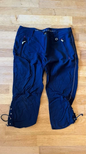 Marinblå 3/4-byxor - Säljer ett par marinblå 3/4-byxor med dragkedjor längs sidorna och snörning vid bensluten. Byxorna har två framfickor med dragkedja och detaljer i rött. Perfekta för en aktiv stil och tillverkade i ett lätt, slitstarkt material. Ingår bälte.