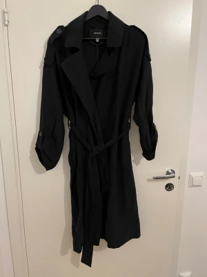 Svart trenchcoat från Vero Moda - Stilren svart trenchcoat från Vero Moda med klassisk krage, axelklaffar och knytskärp i midjan. Jackan har långa ärmar med knappdetaljer och är tillverkad i ett mjukt tyg som ger en snygg siluett. Perfekt för dig som gillar minimalistisk och tidlös stil.