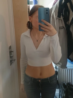 Vit croppad långärmad polotopp - Säljer en vit croppad polotopp med lång ärm och djup v-ringning. Toppen har en klassisk krage och är ribbad i materialet vilket ger en snygg och trendig look. Perfekt att matcha med höga jeans eller kjol.