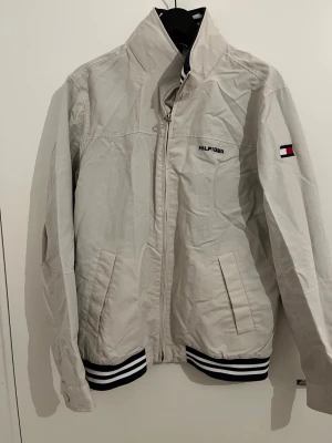 Beige vindjacka från Tommy Hilfiger - Snygg beige vindjacka från Tommy Hilfiger med dragkedja, hög krage och två fickor framtill. Jackan har marinblå och vita ränder på muddar och krage samt Hilfiger-logga på bröstet och ärmen. Perfekt för en sportig och stilren look.