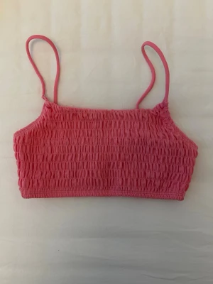 Rosa smockad bikiniöverdel från Nelly Beach - Rosa bikiniöverdel med smala axelband och smockad struktur från Nelly Beach i storlek S. Modellen är rak och enkel, perfekt för stranddagar. Materialet känns mjukt och följsamt mot huden. 