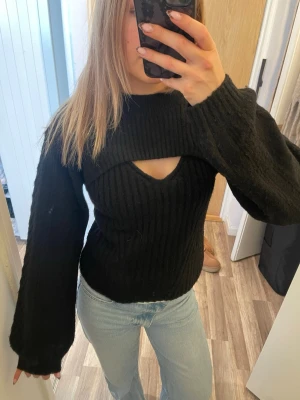 Svart stickad tröja med cut out - Trendig svart stickad tröja med ribbad struktur och unik cut out-detalj vid bröstet. Tröjan har långa, vida ärmar och en hög halsringning. Perfekt för dig som vill ha en edgy och modern look.