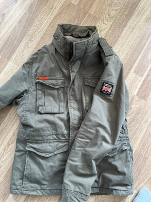 Grön parkajacka från Superdry - Snygg grön parkajacka från Superdry med fyra stora fickor framtill, hög krage och patch med brittisk flagga på ärmen. Jackan har dragkedja och knappar samt en cool militärinspirerad look. Perfekt för dig som gillar streetwear. Är large men skulle säga att den passar medium.