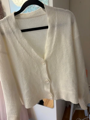 Vit kofta - Mysig vit kofta med v-ringning och tre stora knappar framtill. Koftan har ribbade muddar vid ärmslut och nederkant, samt en fluffig och mjuk känsla. Perfekt för lager på lager och enkel att matcha med olika outfits.