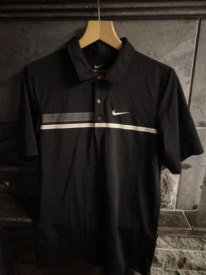 Svart Nike t-tröja Dri-Fit - Svart pikétröja från Nike med Dri-Fit-material, korta ärmar och klassisk krage. Tröjan har vita och gråa ränder över bröstet samt Nike-logga broderad på vänster sida. Perfekt för sportiga tillfällen eller en casual look.🖤💎 För man XXS-S, eller kvinnan S-M. 