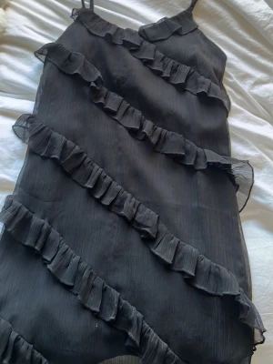 Svart volangklänning Gina Tricot - Säljer en svart kort klänning från Gina Tricot med tunna axelband och flera diagonala volangdetaljer framtill. Klänningen har en lätt och luftig känsla.