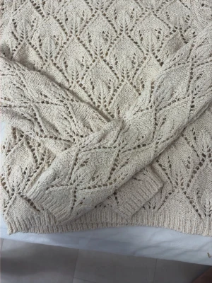 Beige stickad tröja med hålmönster - Säljer en beige stickad tröja med snyggt hålmönster i ett lövliknande motiv. Tröjan har långa ärmar och ribbade muddar vid ärmslut och nederkant. Perfekt för dig som gillar en stilren och mysig look.