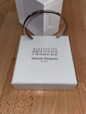 Maison Margiela silverfärgat armband - Snyggt och stilrent armband från Maison Margiela i silverfärgad metall. Armbandet är stelt och har märkets logotyp ingraverad på ovansidan. Perfekt accessoar för dig som gillar minimalistisk och modern design.