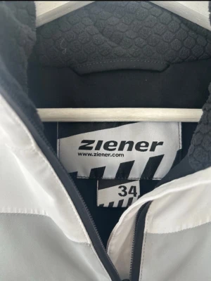 Vit och svart vindjacka från Ziener - Snygg vit vindjacka från Ziener med svarta detaljer och dragkedja framtill. Jackan har ett coolt märke på ärmen och ett mjukt, mönstrat innerfoder. Perfekt för blåsiga dagar och sportiga outfits.