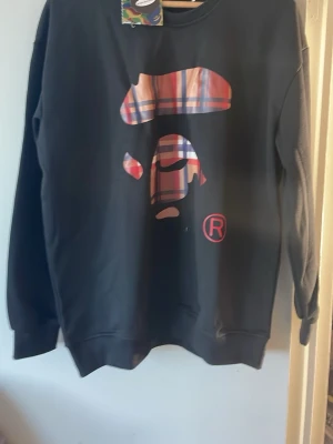 Svart sweatshirt från BAPE - Säljer en svart sweatshirt från BAPE med det klassiska ape-head trycket i röd och blå rutig design på bröstet. På ryggen finns texten 'BY BATHING APE' i samma rutiga mönster. Tröjan har rund hals och ribbade muddar. Perfekt för dig som gillar streetwear. Inget kvitto reserverat 