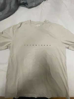 Beige t-shirt från Jack & Jones - Stilren beige t-shirt från Jack & Jones med svart logotyptryck över bröstet. Klassisk rund halsringning och korta ärmar. Perfekt basplagg till garderoben och enkel att matcha med olika stilar.