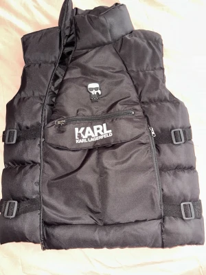Svart dunväst från Karl Lagerfeld - Snygg svart dunväst från Karl Lagerfeld med stor ficka framtill, dragkedja och justerbara spännen i sidorna. Västen har en ikonisk Karl Lagerfeld-logga och en cool patch med Karl-ansikte på bröstet. Perfekt för lager på lager. Som ni ser på sista sidan är början av dragkedjan lite sönder men det går ändå att använda dragkedjan.