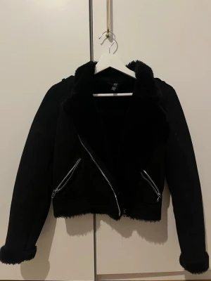 Svart skinnjacka med fuskpälsdetaljer - Kort svart mjuk jacka från H&M med fuskpäls på krage, ärmslut och insida. Jackan har två snedställda dragkedjefickor och coola axelremmar med knappdetaljer. Kortare modell som ger en trendig look och passar perfekt till jeans eller kjol.
