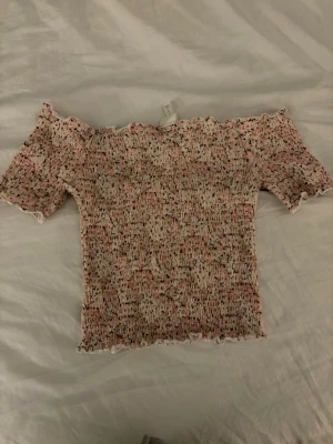 Blommig offshoulder topp i pastell - Söt offshoulder topp med blommigt mönster i rosa, vit och beige. Smockad och stretchig passform med korta ärmar och vågig kant vid ärmslut och nederkant. Säljer då den inte kommer till användning längre! 