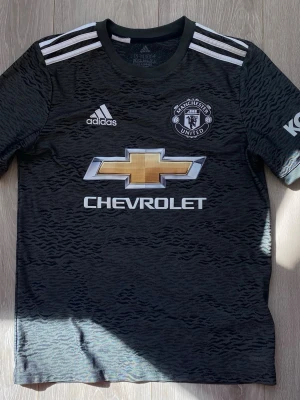 Manchester United tröja - Snygg svart Manchester United bortatröja från Adidas med vita detaljer och mönstrad struktur. Tröjan har klubbmärke, Adidas-logga och Chevrolet som sponsor framtill. Tillverkad i lätt och ventilerande material, perfekt för fotbollsträning eller match. För 13-14 åringar.