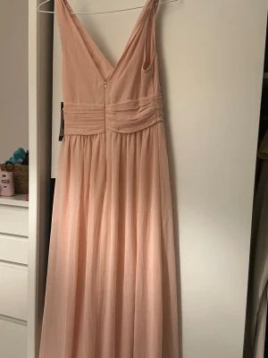 Ljusrosa långklänning med v-ringning - Elegant ljusrosa långklänning med djup v-ringning och breda axelband. Klänningen har markerad midja med veckade detaljer och ett flytande, lätt tyg som ger ett romantiskt intryck. Perfekt för speciella tillfällen.