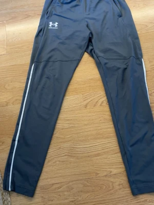 Under armour byxor - Snygga mörkgrå träningsbyxor från Under Armour med vita ränder längs benen. Byxorna har slim passform, resår i midjan och dragkedjor vid bensluten. Perfekta för gymmet eller löprundan. Den ända defekten ser ni på sista bilden därför det låga priset annars är dem i bra skick😊