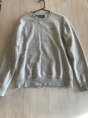 Grå sweatshirt från J.Lindeberg - Snygg grå sweatshirt från J.Lindeberg med diskret logga på bröstet. Tröjan har rund halsringning, ribbade muddar vid ärmslut och nederkant samt en klassisk, avslappnad passform. Perfekt till jeans eller joggers.
