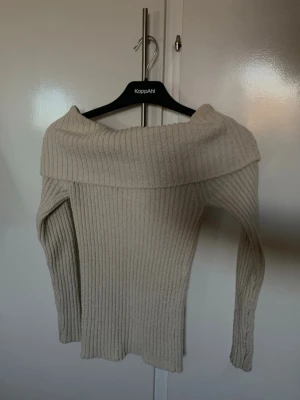 Beige off shoulder stickad tröja - Trendig beige stickad tröja från KappAhl med bred off shoulder-krage och ribbstickad struktur. Långärmad modell i mjukt material som sitter snyggt på kroppen. Perfekt för dig som gillar en stilren och modern look.