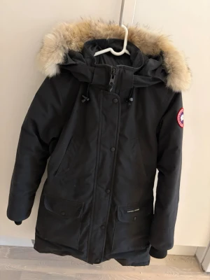 Svart parka Canada Goose - Säljer en svart parkas från Canada Goose med avtagbar huva och äkta päls runt kanten. Jackan har flera fickor framtill, dragkedja och knappar samt den klassiska röda loggan på ärmen. 