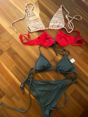 Tre bikinis i olika färger och modeller - Säljer tre snygga bikinis: en randig trekantsbikini i beige och rosa toner med knyt (Swim in peach, S) , en röd bikini med bygel (NELLY, S) och en mörkgrå trekantsbikini med knyt i sidorna (Lager 157, S). Perfekta för sommaren och strandhäng!