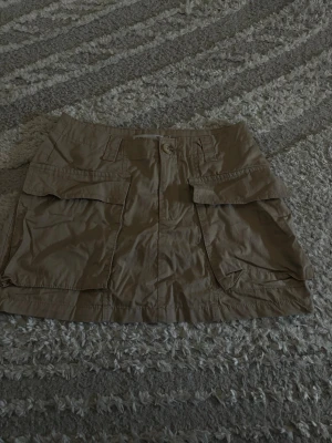 Beige kort kjol - Säljer en beige kort kjol med stora fickor framtill och knappstängning. Skorten har en avslappnad passform och är tillverkad i ett lätt bomullsmaterial. Är 166 cm.