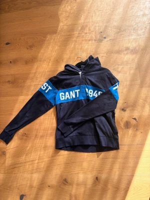 Marinblå hoodie från Gant - Snygg marinblå hoodie från Gant med vit text och blått band över bröstet. Hoodien har huva och långa ärmar. Perfekt för dig som gillar sportig och avslappnad stil. Knappt använd