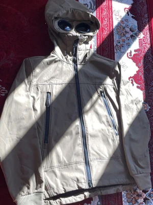 Olivgrön cp company windbraker  - Säljer en olivgrön vindjacka med hel dragkedja, två snedställda fickor med dragkedja och en unik huva med inbyggda glasögon. Jackan har ribbade muddar vid ärmsluten och justerbar huva. Perfekt för dig som vill sticka ut med en cool detalj.