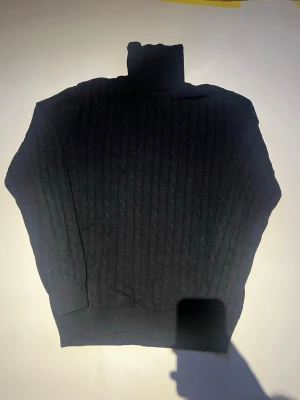 Svart polotröja från John Henric - Kabelstickad svart polotröja från John Henric. Tröjan har hög polokrage, långa ärmar och ribbade muddar. Perfekt och enkel att matcha med olika outfits.