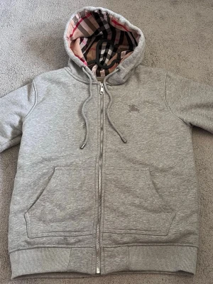 Grå Burberry zip hoodie - Säljer en grå zip hoodie från Burberry med klassiskt rutigt foder i huvan. Tröjan är väldigt sällan använd då den är lite för liten för mig. Då den passar som S-M pris går att diskuteras vid smidig affär. 