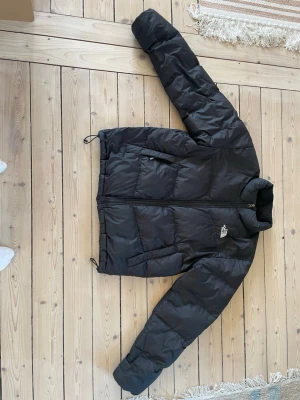 Svart pufferjacka från The North Face - Säljer en svart pufferjacka från The North Face med dragkedja framtill och två sidofickor. Jackan har hög krage, justerbar dragsko i nederkant och klassisk logga på bröstet och ryggen. Perfekt för kalla vinterdagar och riktigt snygg streetstil.