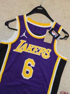 LA Lakers Lebron James, nba swingman jersey. Basketlinne - Snyggt basketlinne från Jordan x NBA med Lakers-tryck och nummer 6. Linnet är lila med gula och vita detaljer, samt svarta sidopaneler. Tillverkat i lätt och ventilerande material, perfekt för basket eller streetwear.
