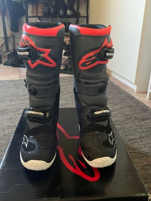 Svarta crossstövlar från Alpinestars - Säljer ett par svarta crossstövlar från Alpinestars med röda detaljer och vita logotyper. Stövlarna har flera spännen för perfekt passform och är tillverkade i slitstarkt syntetmaterial och plast. Perfekta för motocross eller enduro.